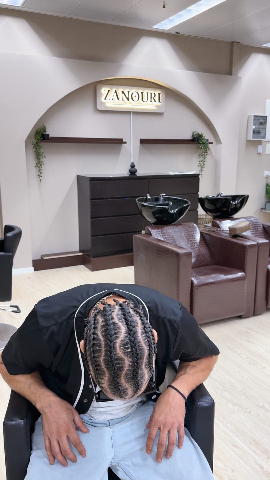 Lange Cornrows in geschwungenen Reihen, Kopf gesenkt, dahinter das ZANOURI-Schild